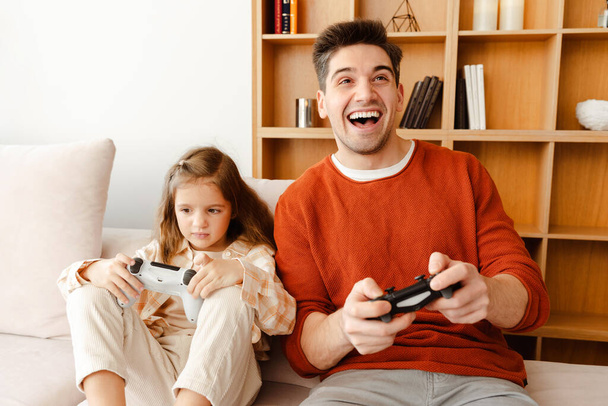 Portrait of positive handsome father and little daughter holding game console, playing video games, sitting in cozy room at home, having fun. Online technology concept - Photo, Image
