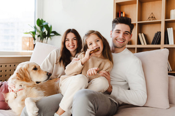 Beautiful smiling family, happy mother, father and cute daughter eating cookies, looking at camera, sitting on comfortable sofa with pet golden retriever. Concept of parenthood, love - Photo, Image