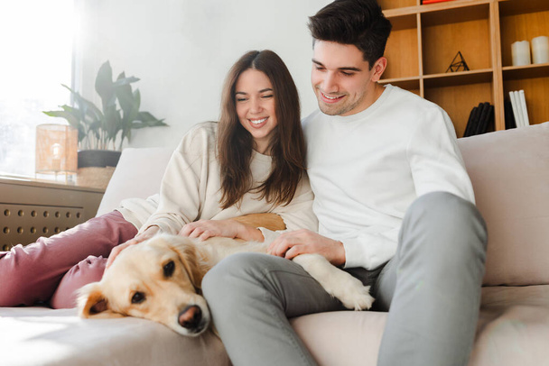 Portrait of smiling young couple, attractive man and woman hugging golden retriever looking at camera while sitting on comfortable sofa in cozy home. Pet care concept - Photo, Image