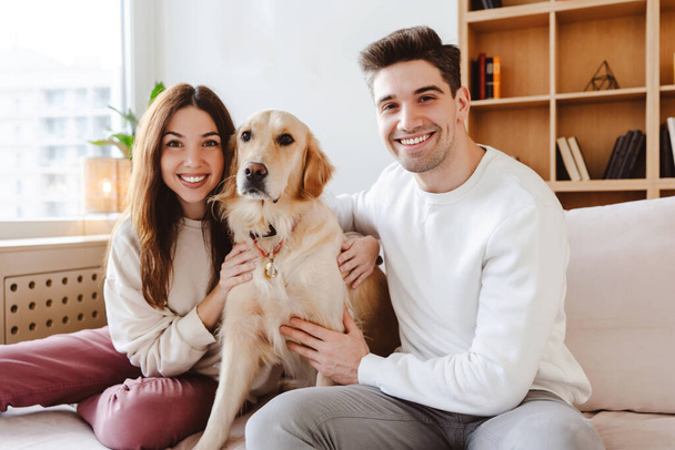 Portrait of positive young couple, attractive man and woman hugging golden retriever looking at camera while sitting on comfortable sofa in cozy home. Concept of love, pet care - Photo, Image