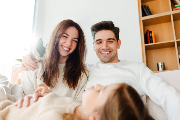 Retrato de sorrir atraente abraço jovem família enquanto sentado em sofá confortável. Pais positivos, juntamente com sua filhinha, se comunicam em uma casa acolhedora. Conceito de amor, paternidade - Foto, Imagem
