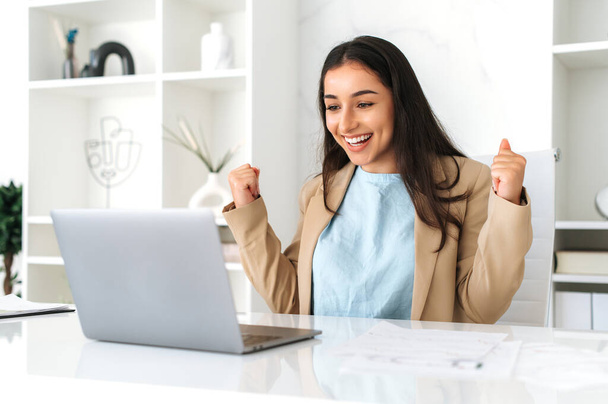 Vrolijke vrolijke Indiase of Arabische zakenvrouw, zit aan een bureau in het kantoor, kijkt graag naar laptop scherm, verheugen promotie op het werk, grote winst, goed werk resultaten, gebaren handen - Foto, afbeelding