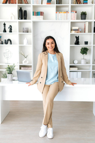 Κάθετη φωτογραφία μιας πανέμορφης επιτυχημένης νεαρής ινδής ή αραβικής επιχειρηματία, γραμματέας, διευθυντής ceo ντυμένος με κομψό κοστούμι στέκεται σε ένα σύγχρονο γραφείο, κοιτάζοντας και χαμογελώντας στην κάμερα - Φωτογραφία, εικόνα