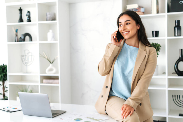 Положительный успех довольно индийская или арабская женщина, сотрудник компании, генеральный директор, сидя на столе в офисе, разговаривая по смартфону, разговаривая с партнером или клиентом, глядя в сторону и улыбаясь - Фото, изображение