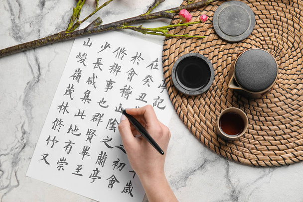 Mano femminile con geroglifici asiatici, penna pennino, inchiostro e tazza di tè su sfondo chiaro. Giornata internazionale della poesia haiku - Foto, immagini