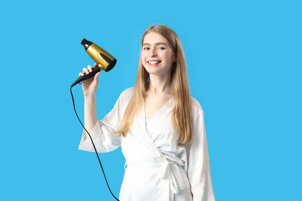 Young blonde woman with hair dryer on blue background - Photo, Image