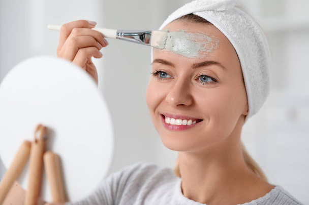 Young woman applying face mask in front of mirror indoors. Spa treatments - Valokuva, kuva