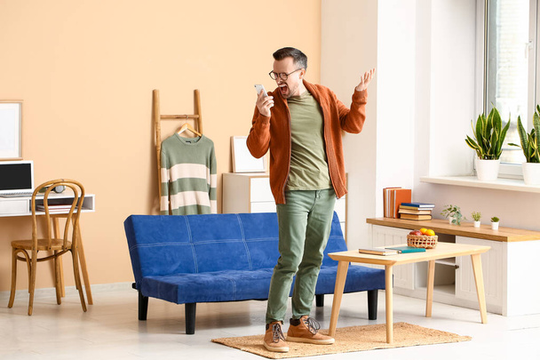 Rozzlobený mladý muž mluví mobilním telefonem v obývacím pokoji - Fotografie, Obrázek