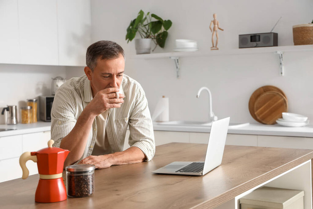 Ώριμος άντρας με φορητό υπολογιστή πίνοντας ζεστό καφέ στο τραπέζι στην κουζίνα - Φωτογραφία, εικόνα