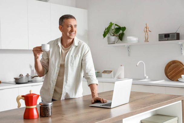 Зрелый человек с чашкой свежего кофе и ноутбука за столом на кухне - Фото, изображение