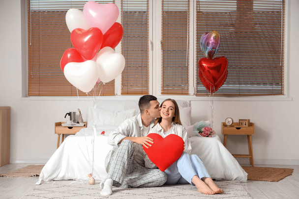 Красивая молодая пара с воздушными шарами в форме сердца, сидящая на полу и празднующая День Святого Валентина дома - Фото, изображение