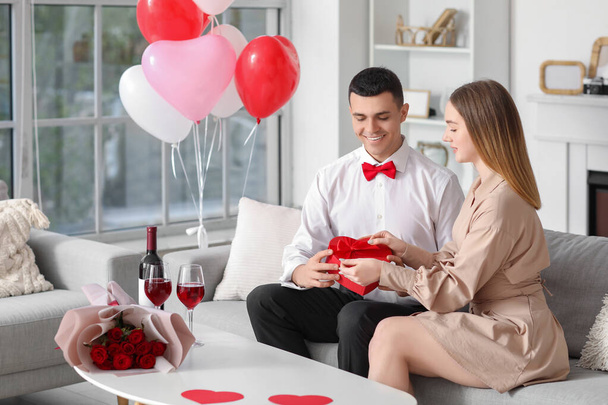 Gyönyörű fiatal pár ajándékdobozzal, csokor virágokkal és borospoharakkal ünneplik a Valentin-napot otthon - Fotó, kép