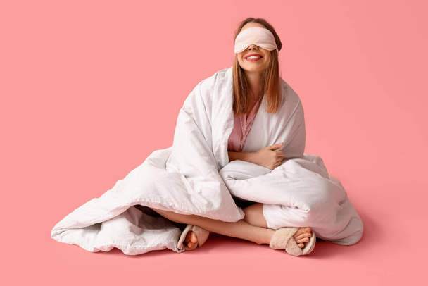 Красивая молодая счастливая женщина с мягким одеялом и сонной маской сидит на розовом фоне - Фото, изображение