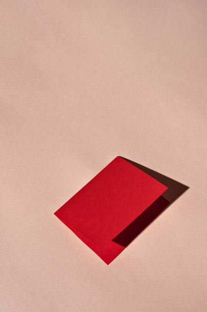Червона складена паперова листівка маскується на світлому персиковому фоні з яскравим світлом і суворими тінями. Мінімальний шаблон брендингу розкішних свят, простір для копіювання. - Фото, зображення