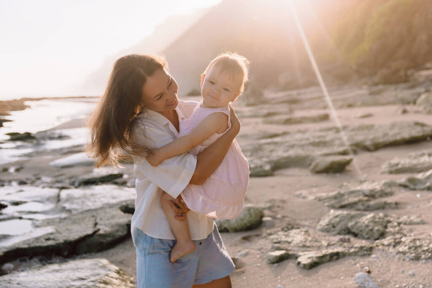Lächelnde Mutter mit kleiner Tochter am Strand bei Sonnenuntergang. Glückliche Familie spaziert an der Küste - Foto, Bild