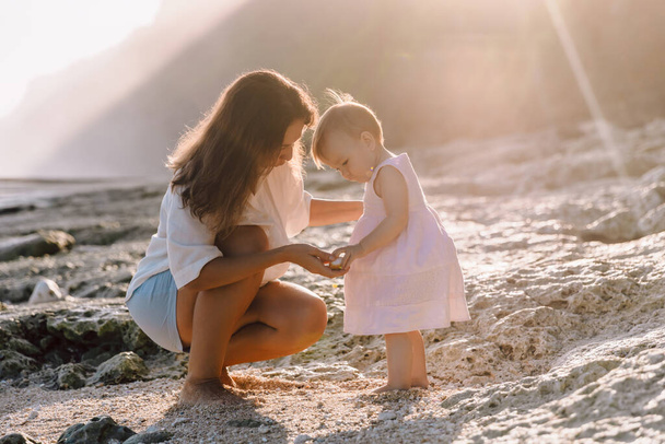 Wesoła matka uczy dziecko chodzić po plaży z odcieniami słońca. Piękna rodzina na wybrzeżu - Zdjęcie, obraz