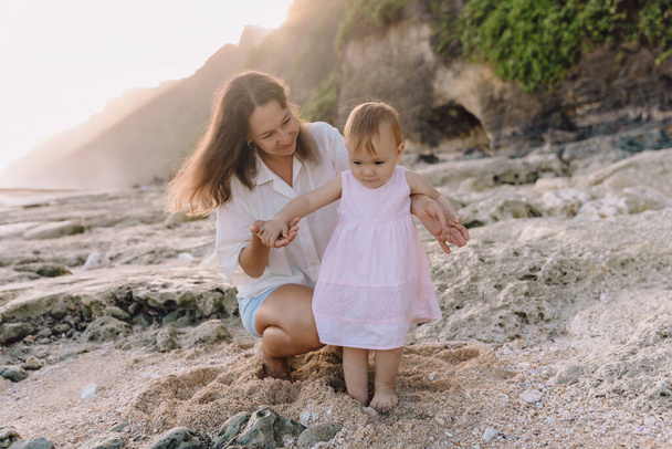 Iloinen äiti opettaa lapselle kävellä rannalla auringonlaskun sävyillä. Kaunis perhe rannikolla - Valokuva, kuva