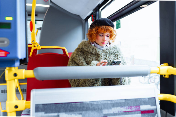 retrato de mujer joven con maquillaje brillante, manicura, vestida, con boina, abrigo de piel verde, usando el teléfono, en el autobús - Foto, imagen