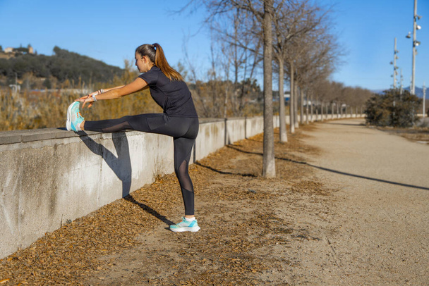 Young runner girl, dressed in tight black sports clothing and ponytail, touching her foot with her hands, stretching her leg leaning on a wall on a path in a public park. - Photo, Image