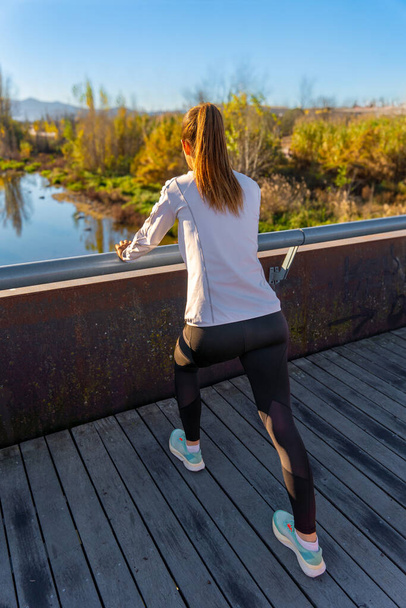 Aantrekkelijk jong runner meisje, met haar haar gebonden in een paardenstaart, het dragen van zwarte panty en een witte sportjas, strekken haar benen kijken naar een rivier over een houten brug in het licht van de dageraad. - Foto, afbeelding