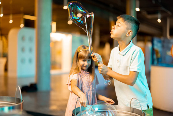 Маленька дівчинка і хлопчик роблять мильні бульбашки, граючи разом і розважаючись у науковому музеї. Концепція дитячих розваг та навчання - Фото, зображення
