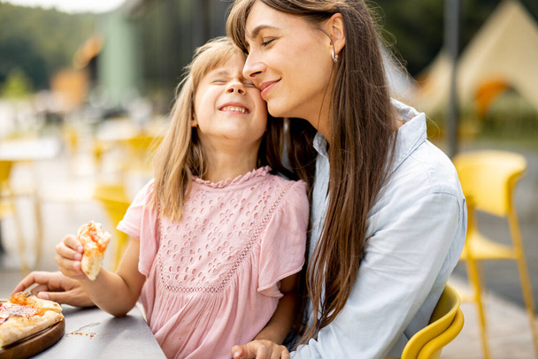 Веселая мама с маленькой дочкой, которая ест пиццу на открытом воздухе, посещает парк развлечений во время летних каникул - Фото, изображение