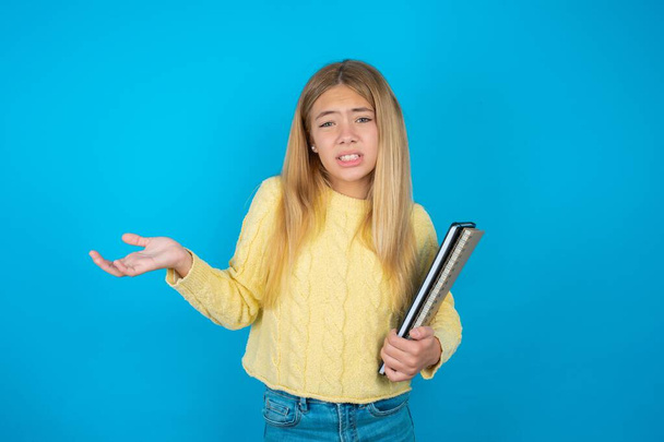 Clueless beautiful kid girl wearing yellow sweater over blue background shrugs shoulders with hesitation, faces doubtful situation, spreads palms, Hard decision - Photo, Image