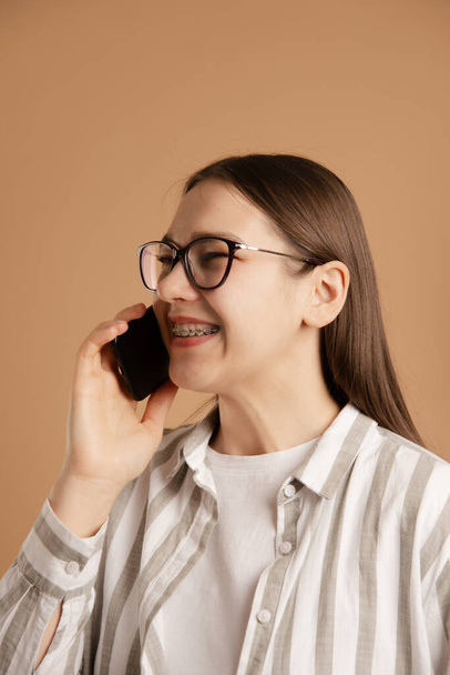 Nuori nainen hammasraudat soittaa älypuhelimeen beige tausta - Valokuva, kuva