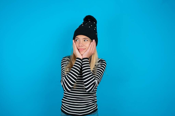 Εμπνευσμένο όμορφο κορίτσι παιδί φορώντας πλεκτό μαύρο καπέλο και ριγέ ζιβάγκο πάνω από το μπλε φόντο κοιτάζοντας copyspace έχοντας σκέψεις για μελλοντικά γεγονότα - Φωτογραφία, εικόνα