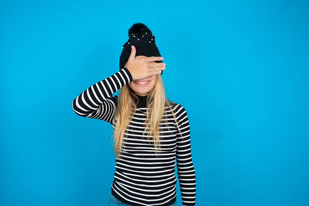 όμορφο κορίτσι παιδί φορώντας πλεκτό μαύρο καπέλο και ριγέ ζιβάγκο πάνω από το μπλε φόντο χαμογελώντας και γελώντας με το χέρι στο πρόσωπο που καλύπτει τα μάτια για έκπληξη. Τυφλή έννοια. - Φωτογραφία, εικόνα