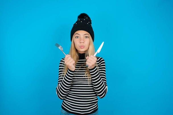 hungry beautiful kid girl wearing knitted black hat and striped turtleneck over blue background holding in hand fork knife want tasty yummy pizza pie - Photo, Image