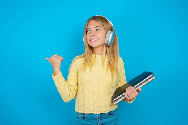 hermosa niña usando suéter amarillo sobre fondo azul escucha pista de audio a través de auriculares inalámbricos puntos pulgar lejos anuncia espacio de copia - Foto, Imagen