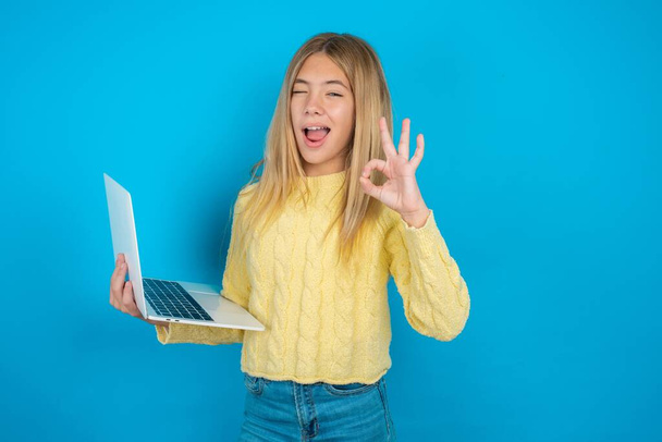 Attraente allegro abile bella ragazza bambino indossa maglione giallo su sfondo blu utilizzando computer portatile che mostra ok-segno winkin - Foto, immagini