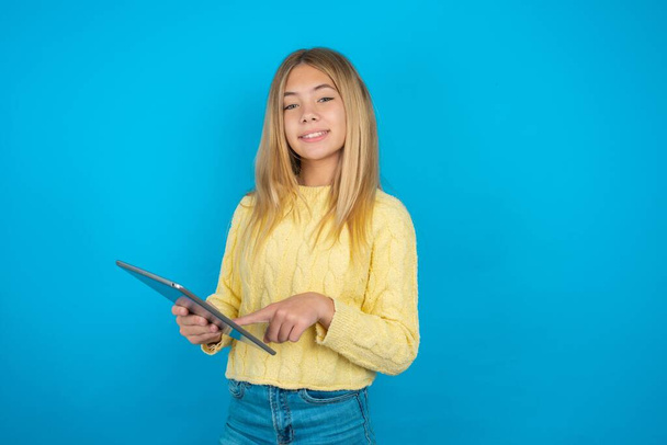 Foto di felice allegro intelligente bella ragazza bambino indossa maglione giallo su sfondo blu tenere tablet navigazione internet - Foto, immagini