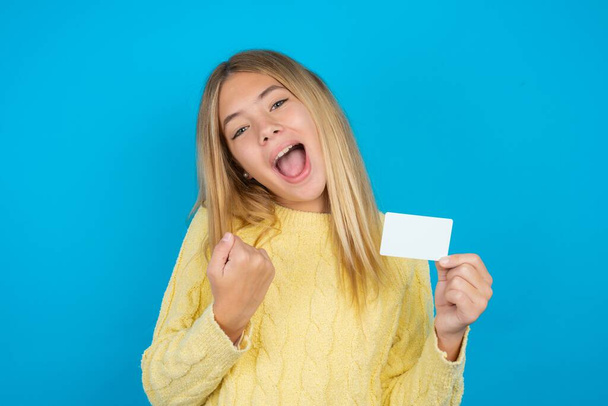 Excited happy positive cheerful smiling beautiful kid girl wearing yellow sweater over blue background hold credit card raise fist in victory - Photo, Image