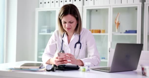 Beautiful therapist consults in clinic using smartphone application. Smiling female doctor holding mobile technology device sending text message and consulting remote patient online - Footage, Video