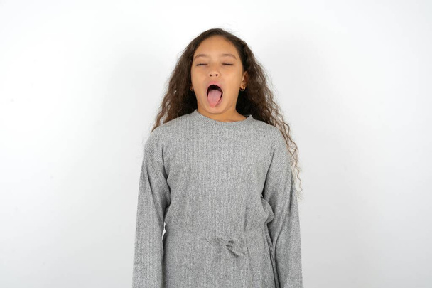 Menina adolescente vestindo vestido cinza contra fundo branco furando a língua para fora feliz com expressão engraçada. Conceito de emoção. - Foto, Imagem