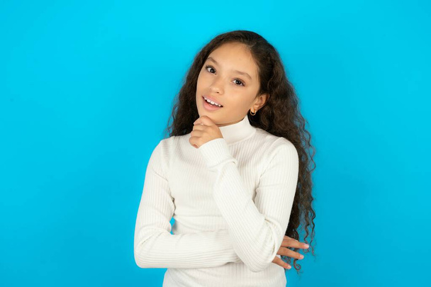 tiener meisje dragen witte trui lacht gelukkig houdt hand op kin uitdrukt positieve emoties glimlacht breed heeft zorgeloze uitdrukking - Foto, afbeelding