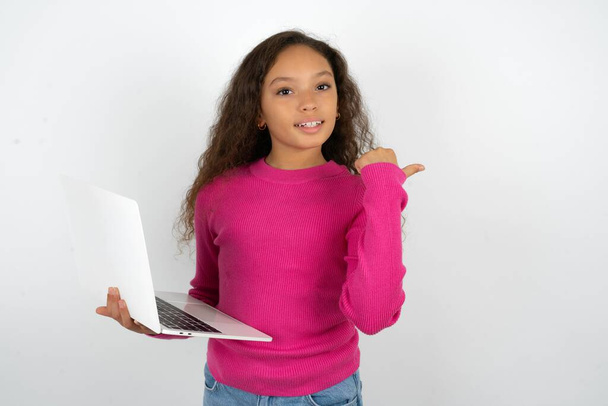 空のスペースを指示するピンクのセーターを身に着けているティーンエイジャーの少女はラップトップを保持します - 写真・画像