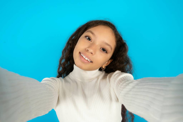 Φωτογραφία του νεαρού όμορφη έφηβη κοπέλα φορώντας λευκό ζιβάγκο πάνω από το μπλε φόντο κάνει selfie - Φωτογραφία, εικόνα