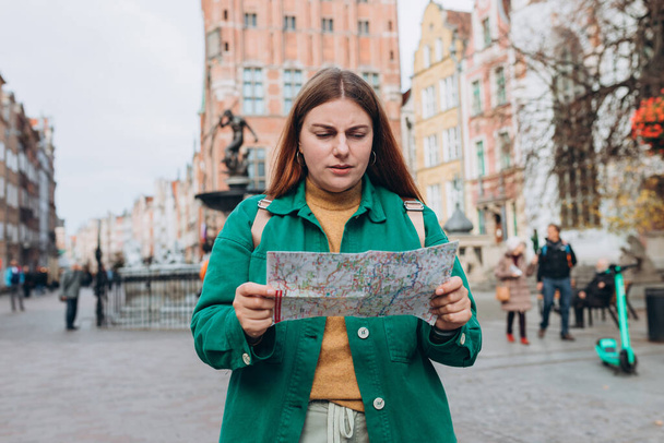 市街地の紙の地図を持つ美しい女性の肖像画. 若い女性旅行者が地図を使って街で迷子になったことを心配した. 旅行する興味深い場所を探索することによるバケーションコンセプト. - 写真・画像