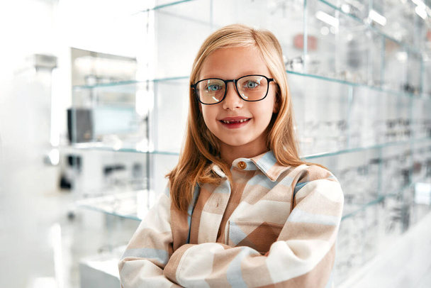 Aranyos lány gyermek optikai szemüvegben áll egy profi boltban. Látásjavítás és szemészet. - Fotó, kép