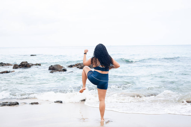 Mulher fitness, de pé, fazendo exercícios de perna na areia da praia com as costas para a câmera. Estilo de vida saudável. - Foto, Imagem