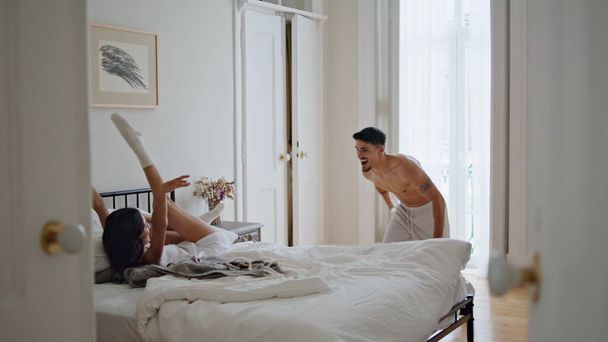 幸せなボーイフレンドはベッドに横たわる妻をジャンプします. 週末のアパートで大声で笑うケアフリースイート. ベッドルームのインテリアで楽しいパジャマカップル. 朝のアクティビティコンセプト - 写真・画像