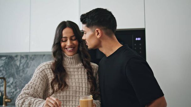 Lächelnde Liebende küssen sich in der morgendlichen Küche aus nächster Nähe. Glückliches Paar kocht zusammen zu Hause. Sexy Ehepartner lachen über modernes Interieur. Verliebte Männer und Frauen amüsieren sich im gemütlichen Haus mit Erdnussbutter  - Foto, Bild