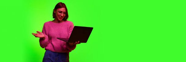 Beautiful young concentrated, positive business woman wearing knitted sweater using laptop while standing against gradient green background. Concept of business, working and studding online. Ad - 写真・画像