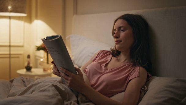Расслабленная девушка читает книгу в постели крупным планом. Расслабленная женщина наслаждается романом перед сном в уютном номере отеля. Бессонная студентка изучает литературу поздно ночью в свете лампы. Концепция отдыха во время сна - Фото, изображение