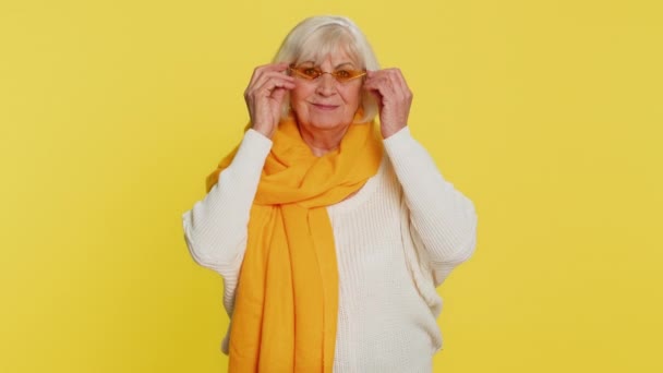Portret van een verleidelijke, stijlvolle oude vrouw met een oranje zonnebril, charmante glimlach, een overwinning vieren, goed positief nieuws. Gelukkig oudere oma gepensioneerde op gele achtergrond - Video
