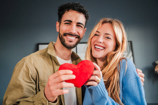 Mladý rozkošný pár s červeným srdcem se usmívá a dívá se na fotoaparát se šťastným výrazem. Přítelkyně a přítel slaví Valentýna slavnost nebo vztah výročí spolu - Fotografie, Obrázek