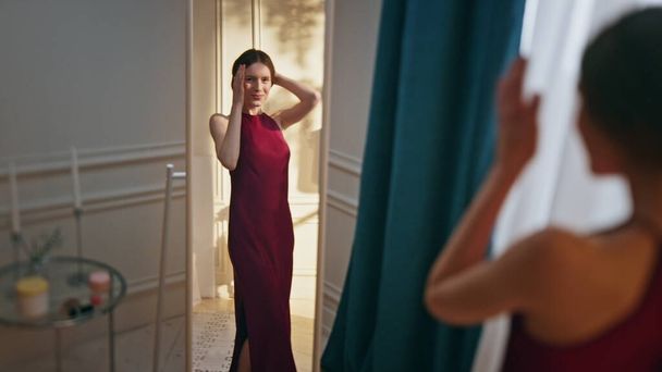Woman try party dress planning outfit in reflection. Smiling sexy girl getting ready for celebration touching hair in sunny room. Relaxed attractive model admiring beauty look mirror at home evening - Photo, Image